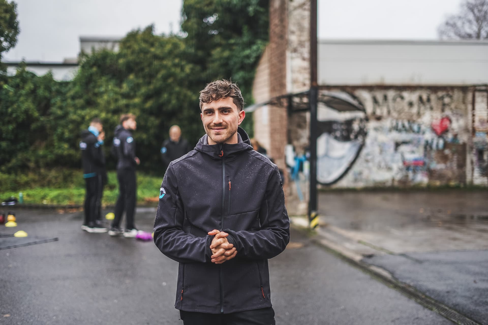 Trainer Mica Oppmann steht in einer schwarzen Jacke in der Natur und lächelt mit gefalteten Händen in die Kamera