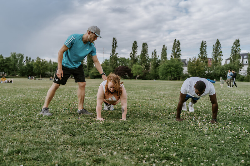Trainer begleitet Teilnehmende bei Liegestützen im Park – Zirkeltraining zuhause mit professioneller Anleitung von Original Bootcamp.